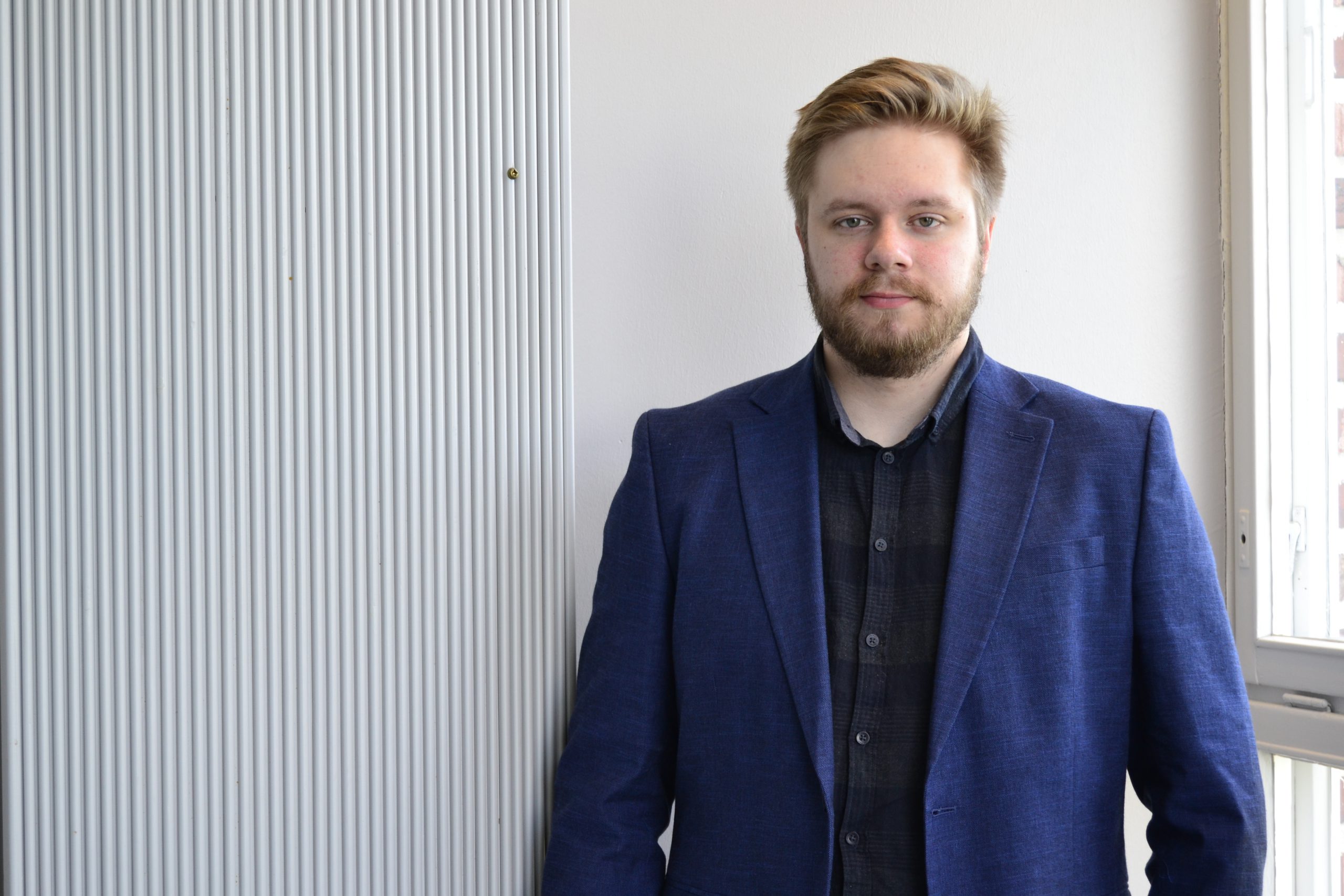Kirjoittaja sinisessä bleiserissä vaaleaa seinää vasten