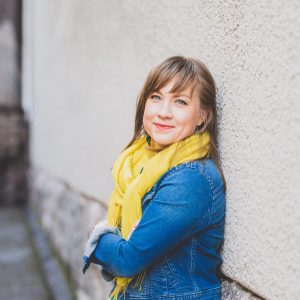 Kuvassa Pauliina Savola. Savola toimi kansainvälisten asioiden asiantuntijana vuosina 2010-2016. Kuvassa Pauliina hymyilee seinään nojaten. Hänellä on kaulassa keltainen huivi ja yllään farkkutakki.